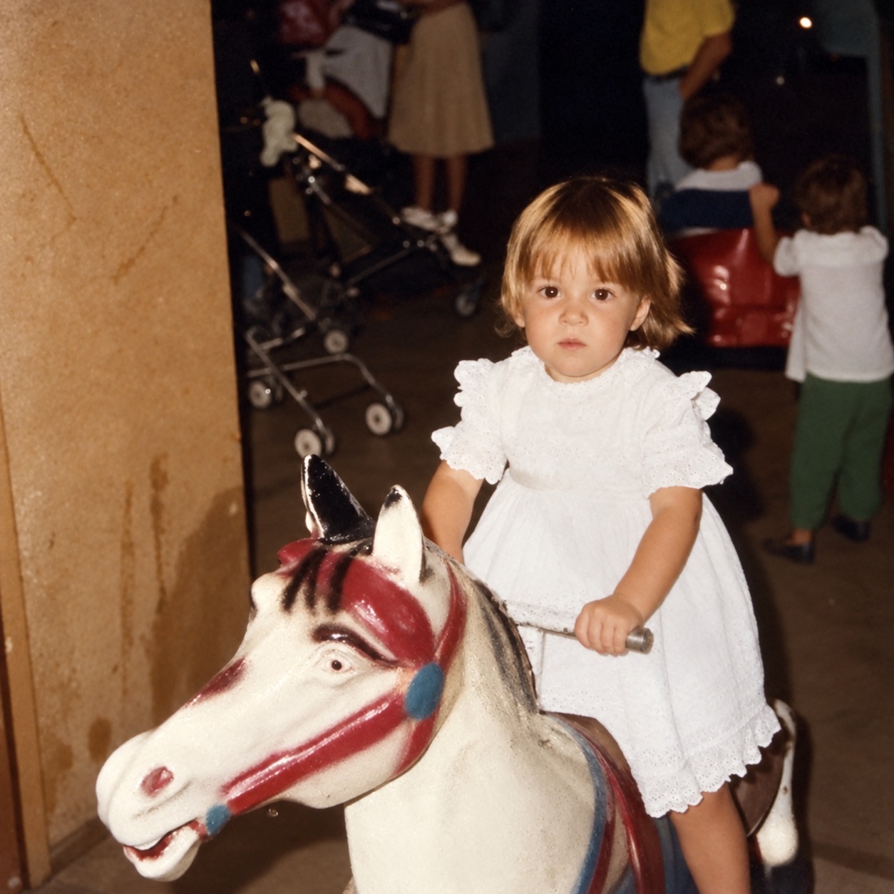 Mili Pérez de niña, con vestido blanco, subida a un poni de juguete pintado de rojo y azul