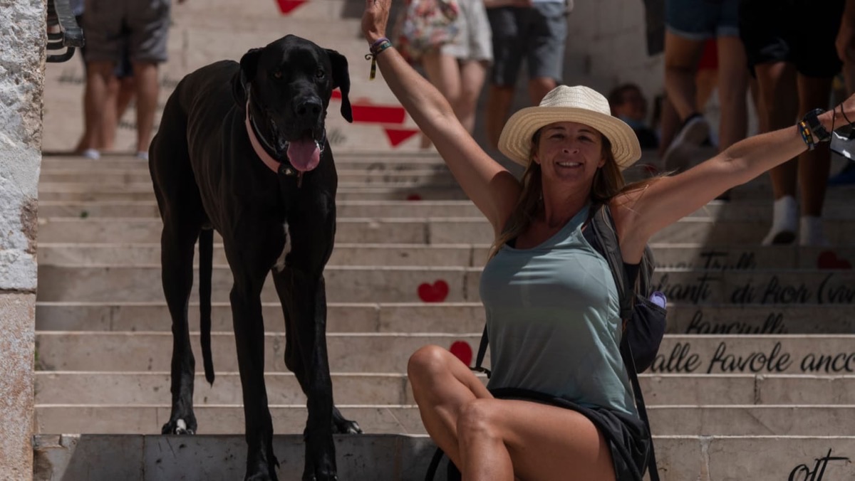Mili Pérez sentada en unas escaleras de piedra con los brazos abiertos, riéndose, junto a su gran danés UX, un perro negro enorme de pie a su lado